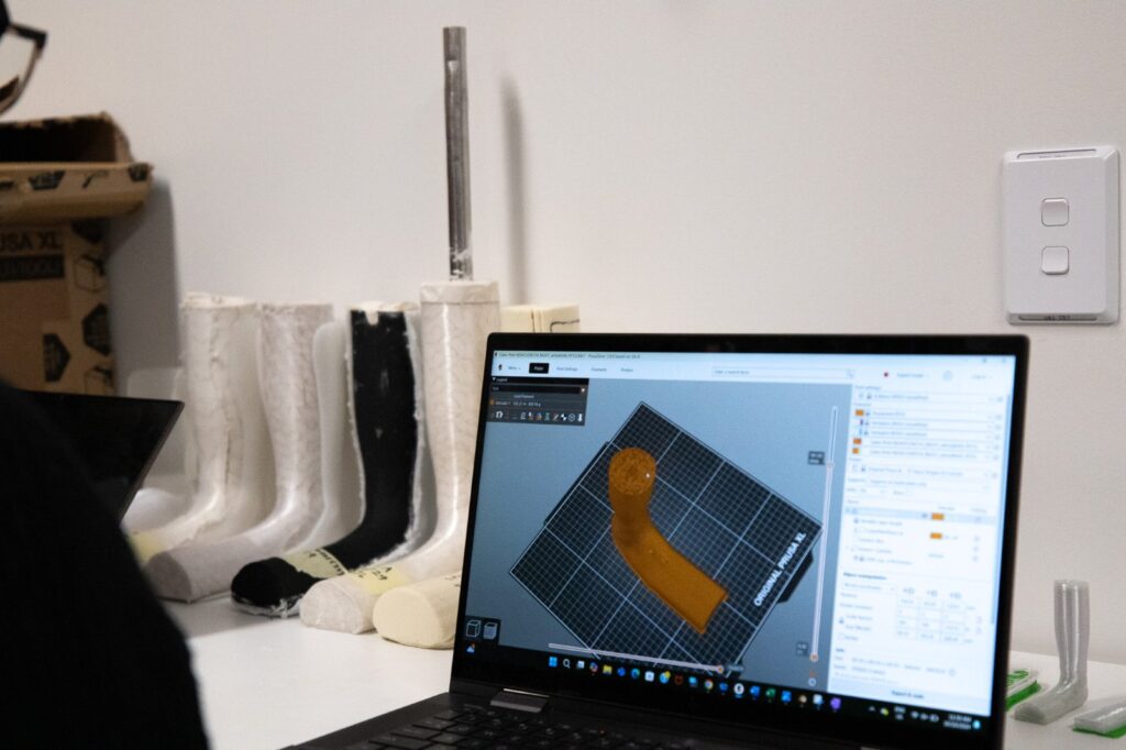 Laptop showing CAD software preparing a custom orthosis for 3D printing, with 3D printed leg moulds behind it