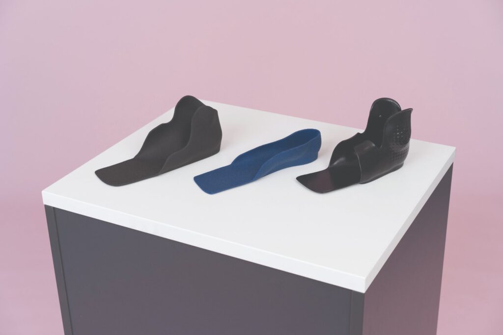 Three custom 3D printed orthoses on a display plinth: a black ankle-foot orthosis, a blue printed foot orthosis and a black supramalleolar orthosis